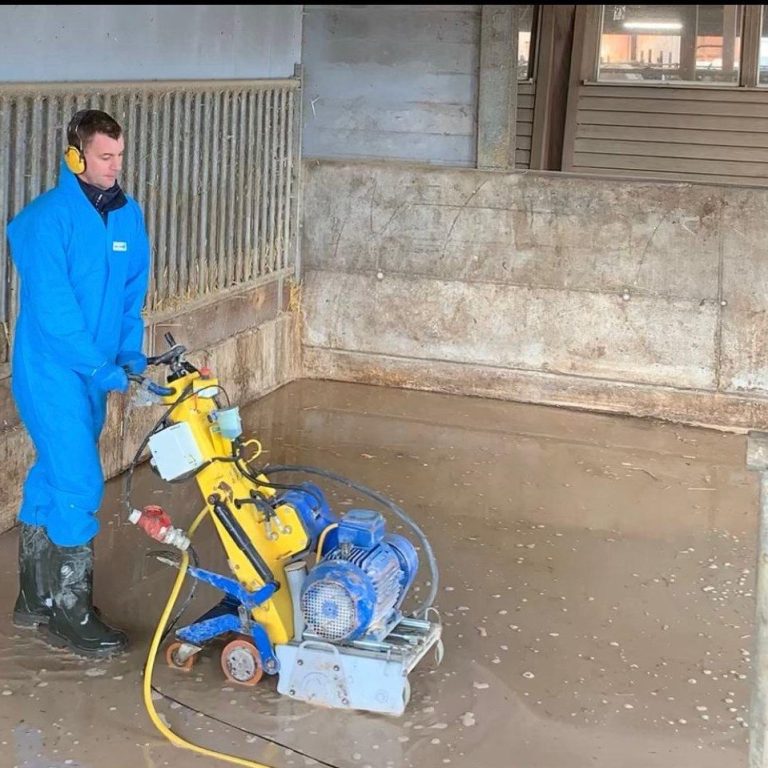 Wir fräsen euren Kuhstall für mehr Trittsicherheit Ein Arbeiter in blauer Arbeitskleidung nutzt eine Maschine zur Bodenbearbeitung in einem Stall.